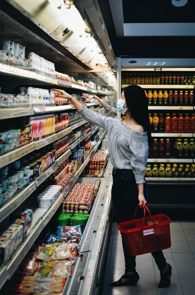 A woman wearing a mask chooses products in a grocery store aisle with a red basket.