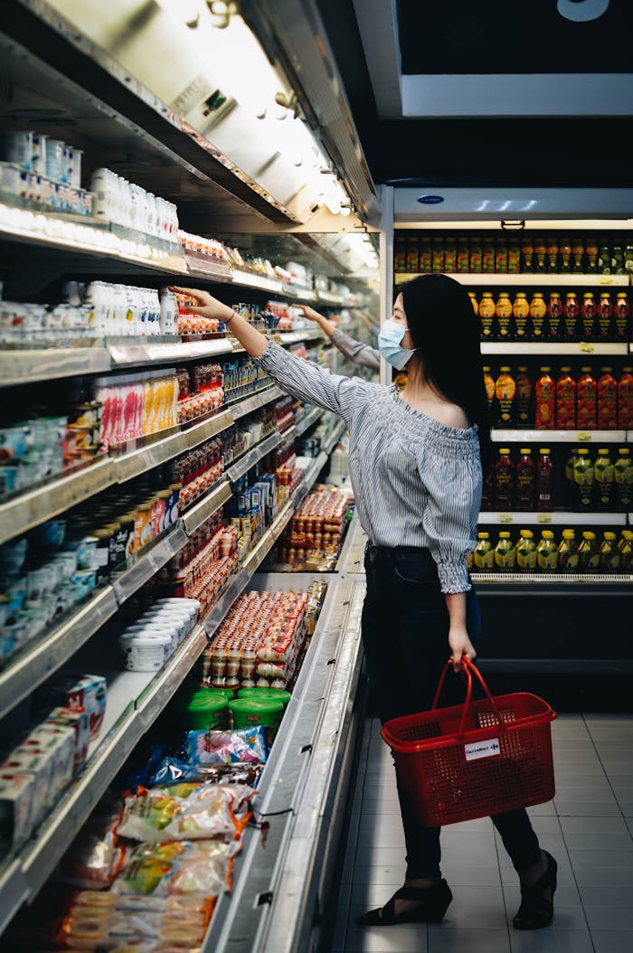 A woman wearing a mask chooses products in a grocery store aisle with a red basket.